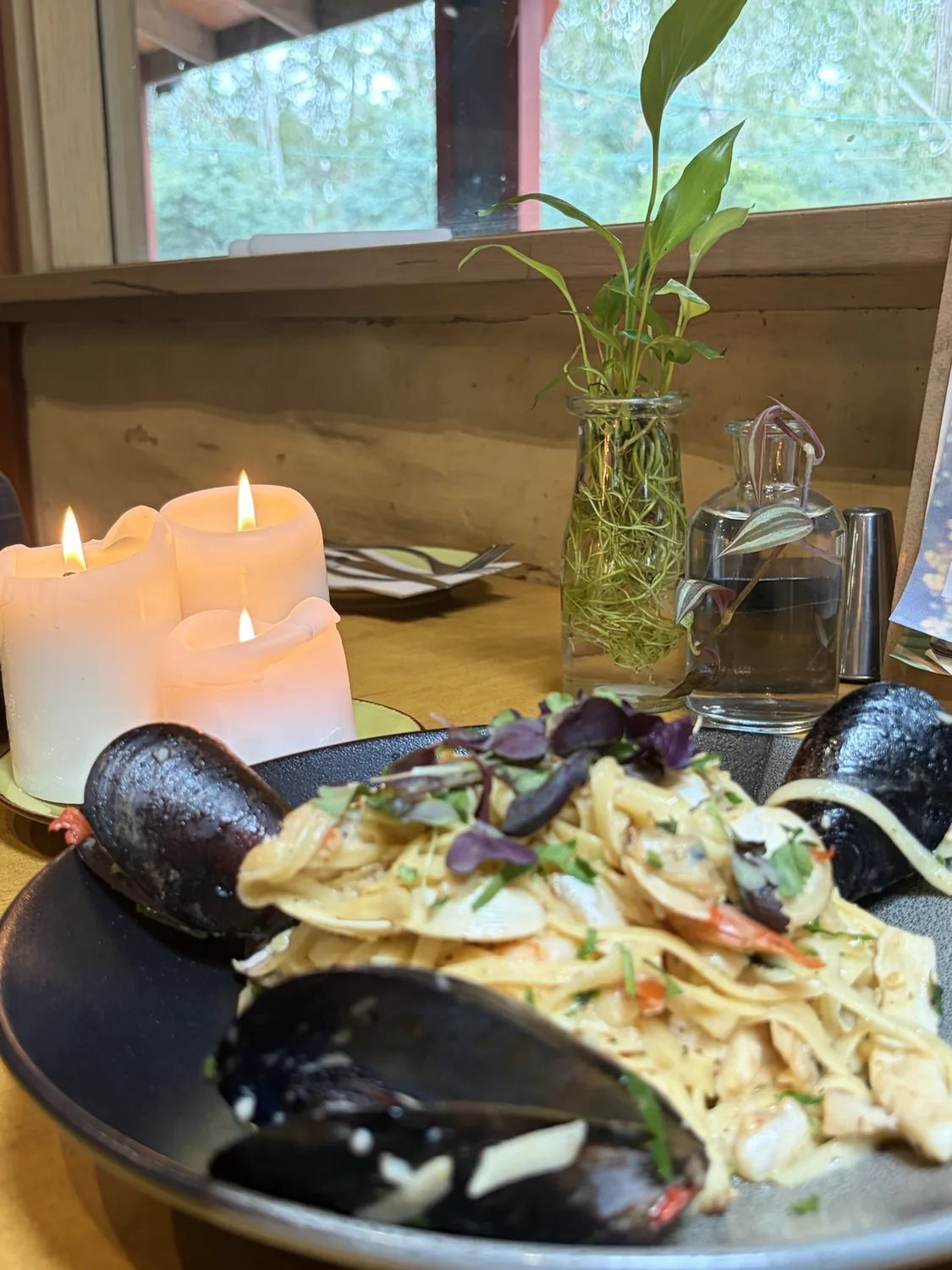 Seafood pasta with mussels in a black bowl, candles and a sprig in a bottle behind, bushland through the window.
